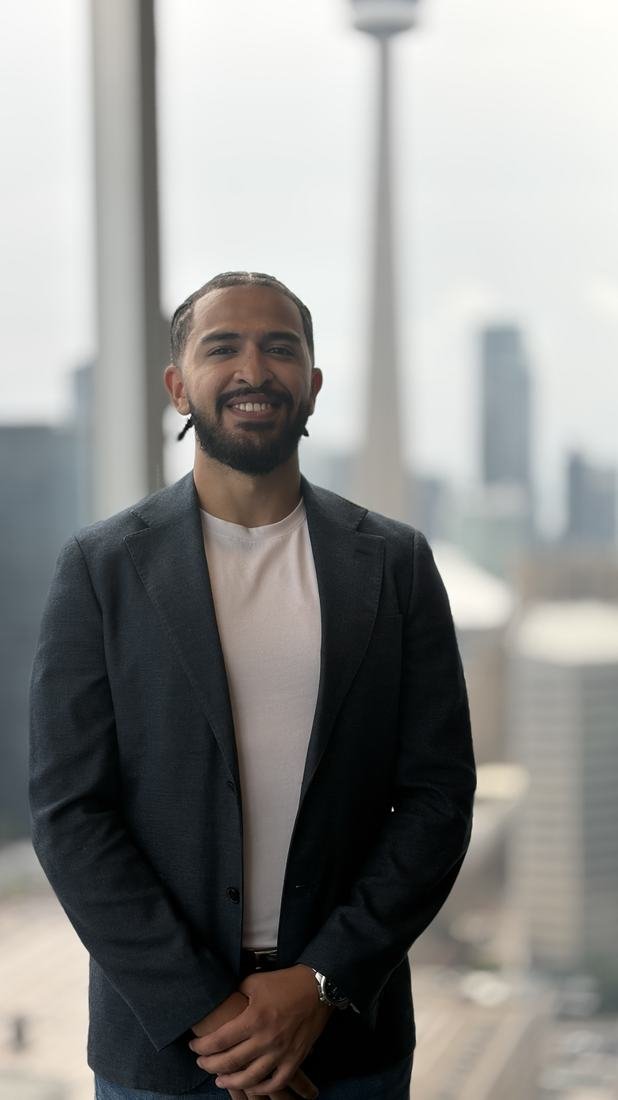 Adnan Sardi in a blazer in a Toronto office with the CN Tower and city skyline behind him